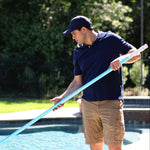 A man in a navy polo shirt, khaki shorts, and a navy cap stands by a swimming pool holding a long light blue pole, with trees and grass in the background.