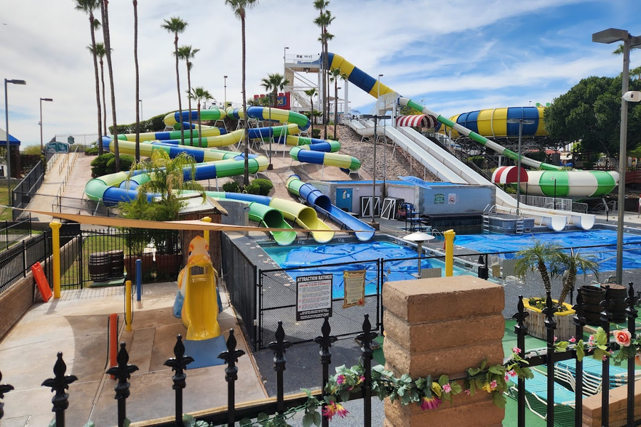 A large outdoor water park with multiple colorful water slides, a swimming pool, palm trees, and a blue sky with clouds in the background.