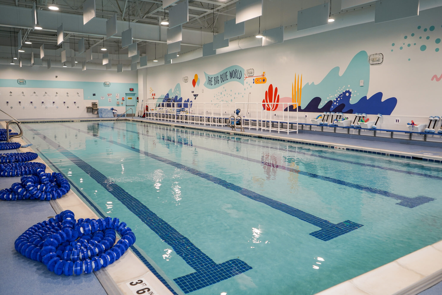 An indoor swimming pool with multiple lanes, blue floating lane dividers on the deck, and a colorful ocean-themed wall mural in the background.