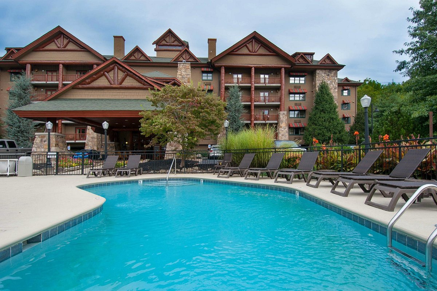 Outdoor swimming pool with blue water in the foreground, black lounge chairs along the poolside, and a large resort-style building with brown wood and stone architecture in the background.