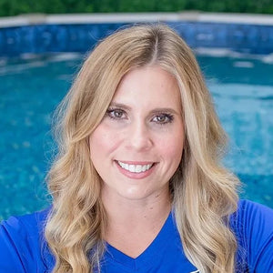 A smiling blonde woman wearing a bright blue long-sleeve baby swim shirt with a green baby fish logo, leaning on a wooden deck next to a swimming pool. A green wall with a 'most mobile BABY FISH' sign is in the background.